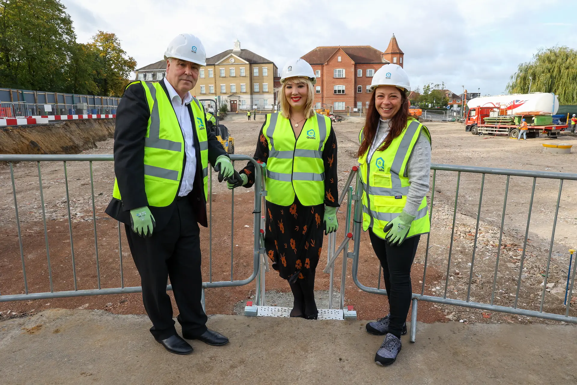 3 people in high visibility vests and hard hats with the Qualis logo on in a construction area smiling at the camera
